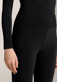 Fekete, elasztikus leggings széles derékrésszel, sima textúrával és testhezálló kialakítással, mellette egy kéz látható, amely a ruházaton pihen.