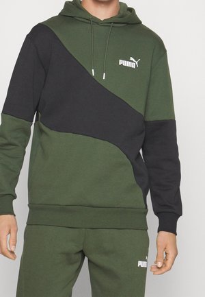 Mann trägt grünen und schwarzen Puma-Hoodie und passende grüne Jogginghose mit Puma-Logos auf Brust und Oberschenkel.