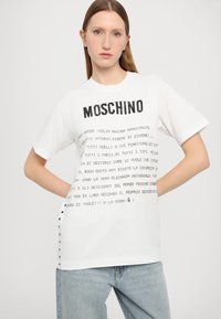 T-shirt de algodão branco com texto preto impresso. Apresenta um decote redondo, mangas curtas e detalhes em botão nas laterais. Nome da marca "Moschino" exibido na parte superior.