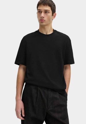 T-shirt nera in cotone con scollo rotondo, maniche corte e vestibilità morbida. Indossata con pantaloni scuri plissettati.