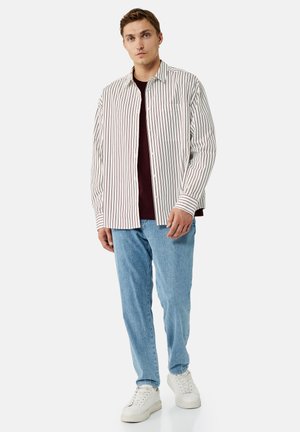 Mann steht und trägt ein weißes Hemd mit roten Streifen, ein bordeauxfarbenes Unterhemd, hellblaue Jeans und weiße Sneakers vor einfarbigem Hintergrund.