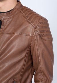 Braune Lederjacke mit einem gesteppten Schulterdesign, glatter Textur und einer Zipptasche auf der unteren linken Seite.
