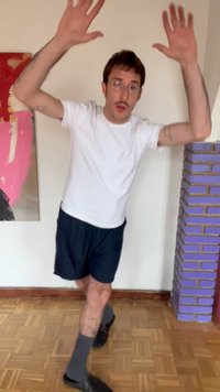 Mężczyzna w białej koszulce i czarnych krótkich spodenkach podnoszący ręce, stojący na jednej nodze na parkiecie obok fioletowej ceglanej kolumny i kolorowej ściennej sztuki.
