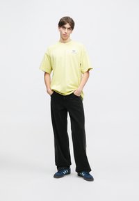 Żółta bawełniana koszulka z okrągłym dekoltem i małym logo na piersi, zestawiona z czarnymi szerokimi spodniami oraz niebieskimi butami z kontrastowymi paskami.