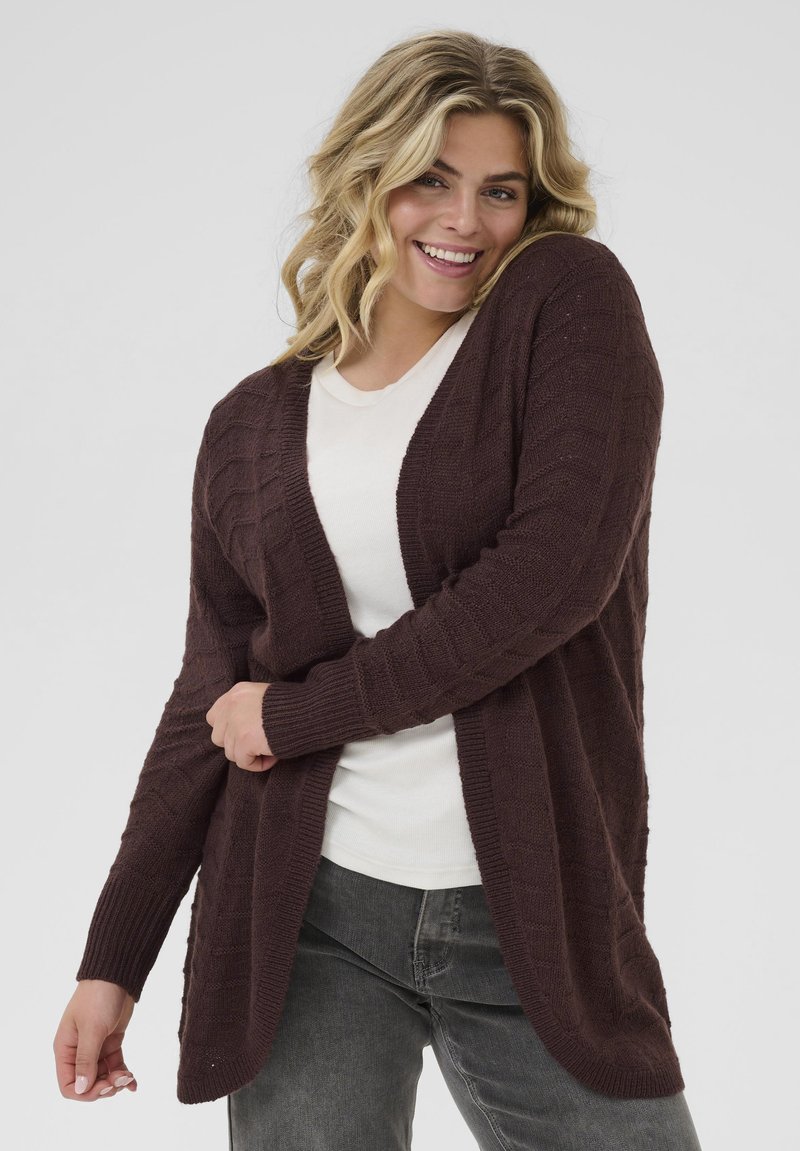 Cardigan maroon tricoté avec un col en V, à ouverture frontale, et des poignets côtelés. Tissu texturé avec un motif géométrique subtil. Stylé par-dessus un haut blanc.