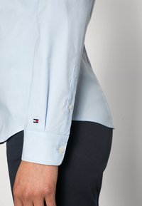 Muñeca de camisa de manga larga azul claro con dos botones y un pequeño logotipo rojo, blanco y azul, llevada con pantalones oscuros.