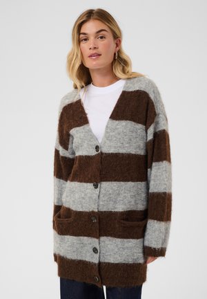 Femme portant un cardigan duveteux à larges rayures marron et gris par-dessus une chemise blanche, debout devant un fond clair uni.