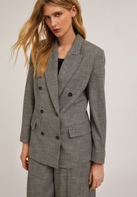 Harmaa houndstooth-mallin kaksirivinen bleiseri mustilla nappeilla, kauluksen etureunalla ja kahdella etutaskulla. Teksturoitu kangas ja muotoiltu istuvuus.