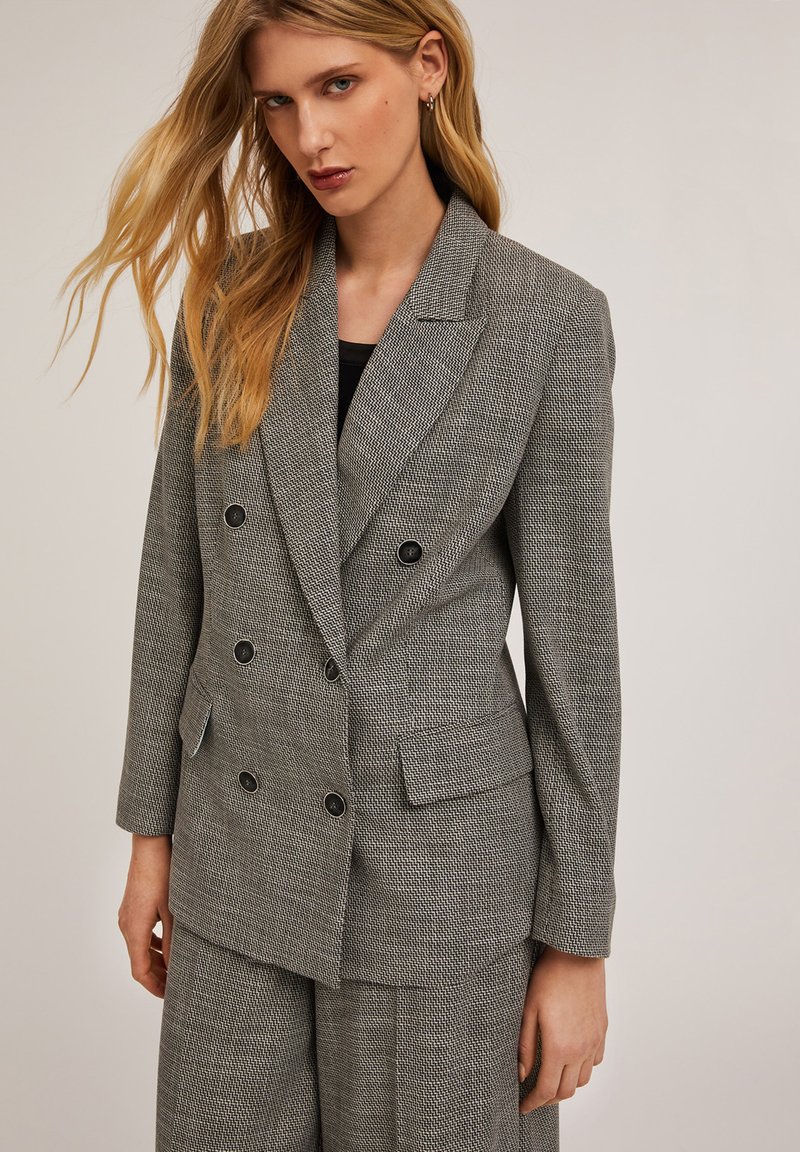 Harmaa houndstooth-mallin kaksirivinen bleiseri mustilla nappeilla, kauluksen etureunalla ja kahdella etutaskulla. Teksturoitu kangas ja muotoiltu istuvuus.
