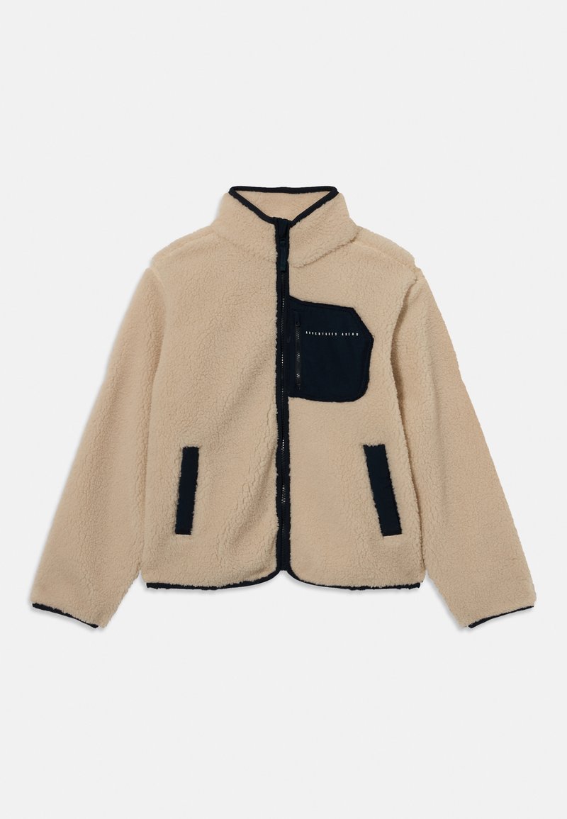 Beige fleece jack met zwarte afwerking, volledige voorrits, twee zijzakken met zwarte afwerking, en een zwarte borstzak met rits en tekst.