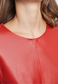 Top en cuir rouge avec un col arrondi et une texture lisse. Présente des coutures discrètes et un doux éclat.