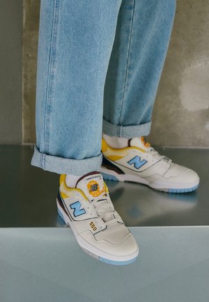 Osoba ubrana w jasnoniebieskie dżinsy i białe sneakersy New Balance 550 z niebieskimi i żółtymi akcentami, stojąca na szarej powierzchni.