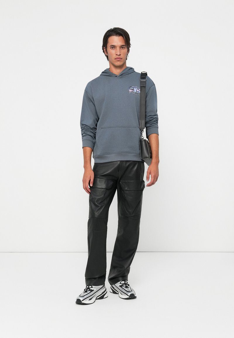 Joven que lleva una sudadera gris, pantalones de cuero negros, zapatillas blancas y un bolso negro de hombro, de pie frente a un fondo blanco sencillo.