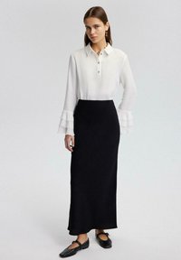 Camisa blanca con botonera y mangas largas con volantes, combinada con una falda negra hasta el suelo. La modelo lleva zapatos planos negros.