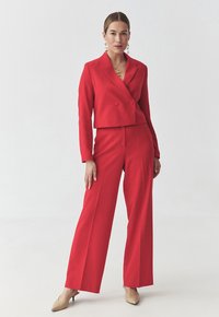 Blazer rosso su misura con doppi bottoni, svasato in vita. Pantaloni a gamba larga coordinati, di texture liscia. Indossato con tacchi beige e accessori.