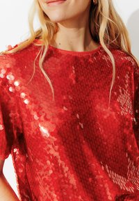 Haut rouge à sequins avec une coupe ample, un col rond et des manches courtes. La surface est ornée de sequins brillants et superposés.