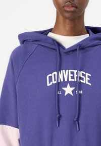 Hoodie roxa feito de tecido suave, apresentando um logo branco "CONVERSE ALL STAR" e cordões, com detalhes em rosa nas mangas.