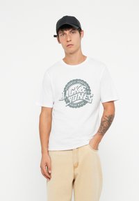 Hvit kortermet t-skjorte med en sirkulær grønn grafikk som leser "JACK & JONES AUTHENTIC JEANSWEAR." Lysfargede, avslappede bukser. Svart caps.