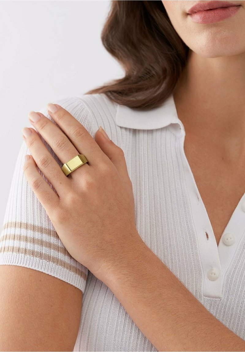 Femme portant une chemise en maille blanche à col avec des rayures beige tient son épaule gauche, montrant une bague épaisse rectangulaire en or à son doigt.