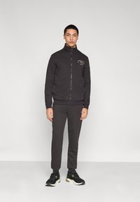 Μαύρο αθλητικό μπουφάν με φερμουάρ και ψηλό γιακά, με λογότυπο στο στήθος. Συνδυάζεται με μαύρο αθλητικό παντελόνι jogging και μαύρα αθλητικά παπούτσια.