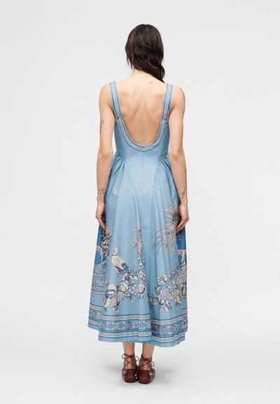 Γυναίκα με τατουάζ φοράει ένα αμάνικο μπλε φόρεμα με βαθιά στρογγυλή πλάτη και σχέδιο με λουλούδια και πουλιά, συνδυασμένο με καφέ παπούτσια με κορδόνια.