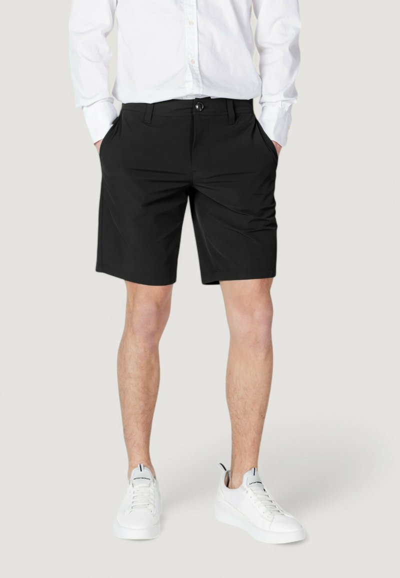 Pantalones cortos negros de tejido ligero, con un corte clásico que incluye dos bolsillos laterales y un cierre de botón. Llevados con zapatillas blancas.