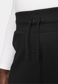 Detailní záběr na černý elastický pás kalhot se šňůrkami proti bílé rukávu košile na těle osoby.