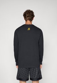 Sort langærmet skjorte med rund halsudskæring, prydet med et lille gult logo på den øverste ryg, parret med mønstrede sorte shorts.