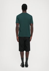 Tumši zaļa kokvilnas T-krekls uz modeļa, kombinēts ar melniem šortiem. Audums izskatās gluds; T-krekliņam ir klasisks apaļš kakls un īsas piedurknes.