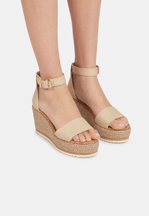 Sandales compensées beige avec brides à la cheville et semelles plateformes tressées portées pieds nus sur un fond uni.