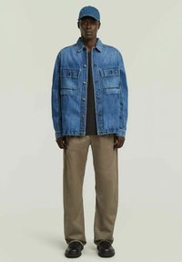 Uomo con giacca di jeans blu, maglietta nera, pantaloni beige, scarpe nere e berretto blu, in piedi contro uno sfondo chiaro e semplice.