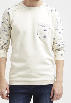 Mand iført offwhite sweatshirt med blå nautiske motiver på ærmer og lomme, kombineret med mørkeblå jeans.