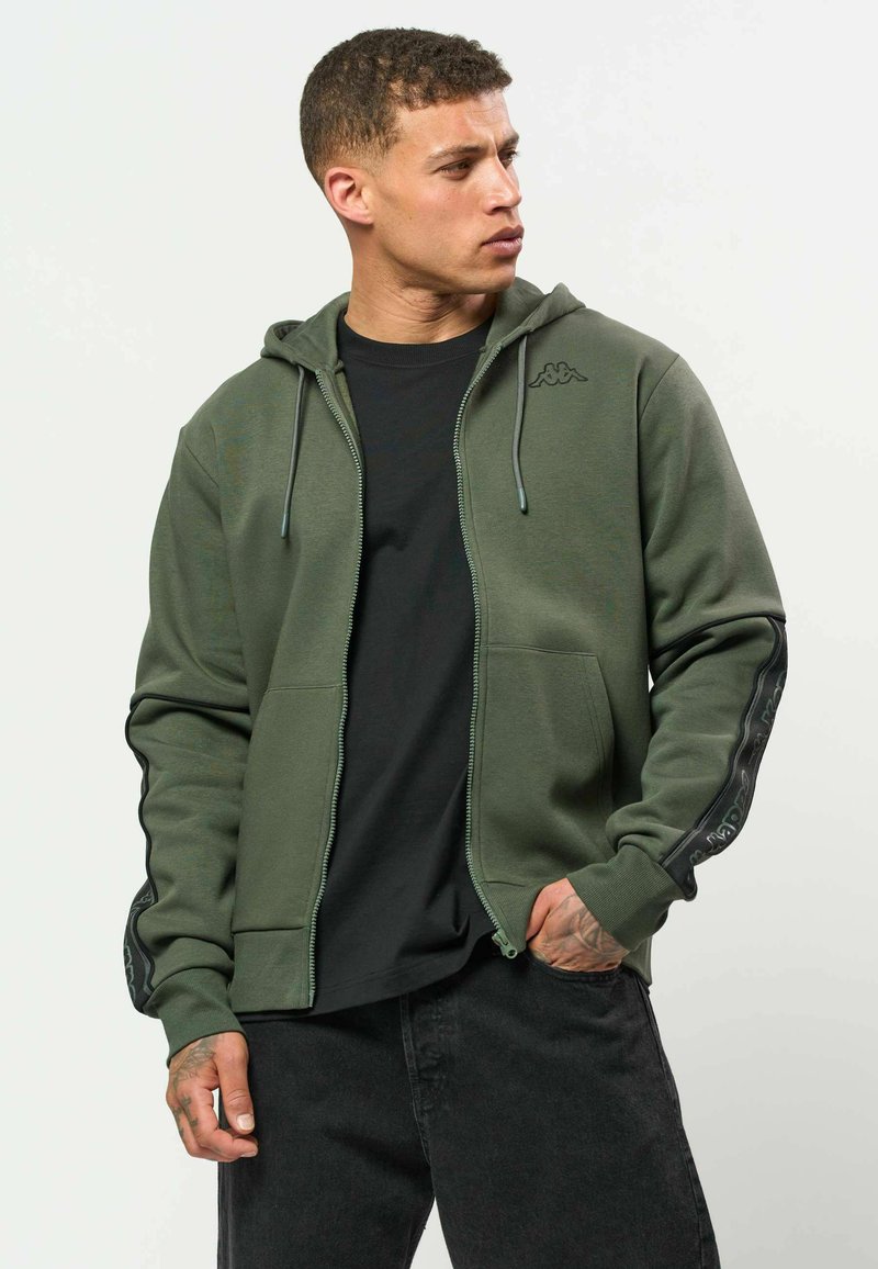 Un jeune homme regarde sur le côté, portant une veste à capuche zippée verte avec des bandes noires sur les manches et un t-shirt noir en dessous.