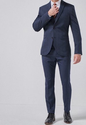 Un homme ajuste une cravate orange brûlé en portant un costume bleu foncé sur mesure et des chaussures habillées noires sur un fond clair uni.