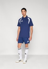 Αθλητική πόλο μπλούζα σκούρου μπλε με λευκές λεπτομέρειες, συνδυασμένη με αντίστοιχα σορτς. Λευκές κάλτσες με μαύρες ρίγες και λευκά ποδοσφαιρικά παπούτσια με μαύρες λεπτομέρειες.