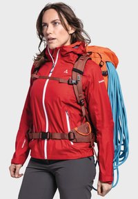 Red waterproof jacket with white zippers, brown harness, and an orange backpack. Blue rope attached. Gray pants complete the outdoor gear.