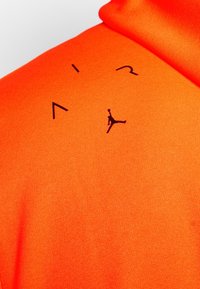 Felpa arancione brillante con un tessuto testurizzato. Presenta loghi neri di "AIR" e una silhouette di una figura che salta nella zona del petto.
