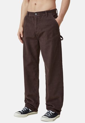 Mann trägt lockere dunkelbraune Cordhosen mit seitlicher Schlaufe und schwarze Low-Top-Sneakers, steht mit einer Hand in der Tasche.