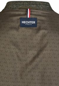 Bruine stof met een herhaaldelijk "H"-patroon, voorzien van een gestructureerde kraag en een marineblauw label met de tekst "HECHTER PARIS" en een rood, wit en blauw lusje.