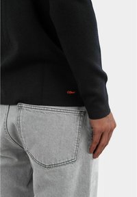 Maglione nero con maniche lunghe, caratterizzato da un piccolo logo rosso vicino all'orlo, abbinato a jeans in denim grigio chiaro che mostrano i dettagli della tasca posteriore.