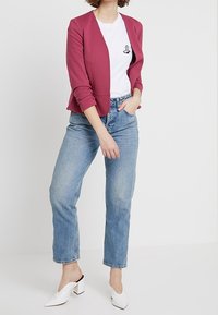 Figurpassad vinröd kavaj över en vit T-shirt med en handgrafik, i kombination med ljusblå raka jeans och vita blockklacksmuler.