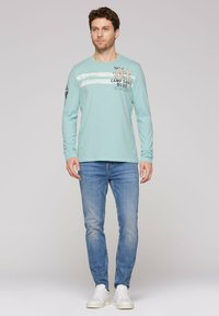 Lange mouw lichtgroene blouse met witte strepen en grafische tekst; gecombineerd met lichtblauwe jeans en witte sneakers. Soepele textuur.