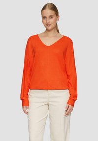 Orange, langärmliger V-Ausschnitt-Pullover aus weichem Material, kombiniert mit hellen Hosen. Der Pullover hat eine lockere Passform und minimale Nähte.