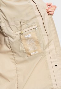 Main tenant une veste beige ouverte montrant une poche intérieure zippée, un cordon réglable et une étiquette beige avec un texte blanc et le nom de la marque Aigle.