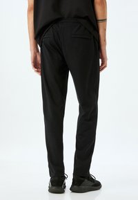 Pantalones de chándal negros con cinturilla elástica, que cuentan con dos bolsillos traseros y una textura de tela suave y lisa. Combinados con zapatos negros.