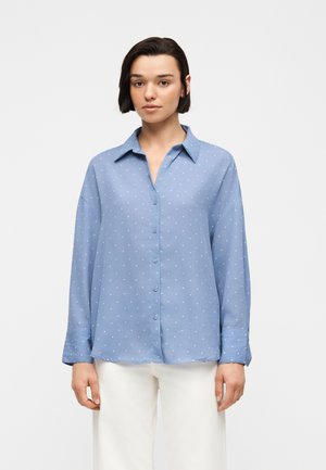 Jeune personne aux cheveux courts et foncés portant une chemise bleue claire à boutons avec des pois blancs et un pantalon blanc sur un fond uni.