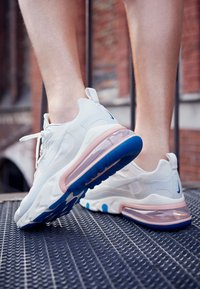 Nohy obuty v bílo-růžových teniskách Nike s modrými podrážkami stojí na kovovém roštu venku u pozadí cihlové zdi.