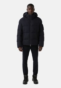 Granatowa puchowa kurtka z kapturem, z zamkiem błyskawicznym z przodu, dwoma bocznymi kieszeniami i teksturowaną tkaniną. Noszona na ciemnych spodniach i czarnych trampkach.