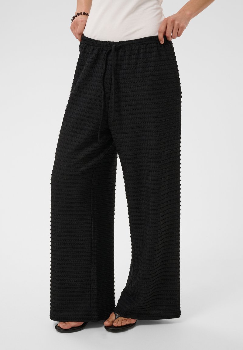 Personne portant un pantalon noir ample texturé à jambes larges avec ceinture à cordon et sandales noires, debout devant un fond uni.
