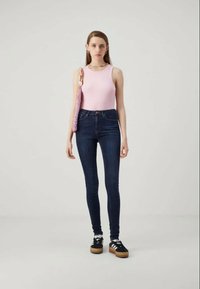 Jovem mulher vestindo uma blusa rosa sem mangas, jeans skinny escuros, ténis plataforma pretos, colar de contas coloridas e carregando uma mala em rede roxa.